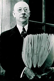 portrait of a Henry Allan Gleason, wearing a suit and glasses, holding three folders of herbarium specimens