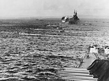 Two cruisers, one in front left foreground,a second in the upper distance is firing. Amphibious vehicles are headed to the right.
