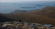 A view from a high rocky eminence with brown moorland below and a vista of small brown islands scattered in the sea beyond. A low bank of fog obscures the horizon.