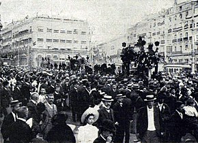 A dense crowd fills the streets of a city; many men wear hats