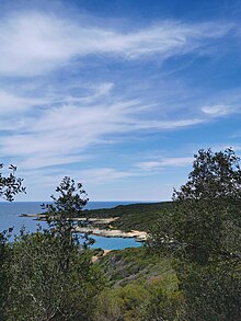 A bright blue sky has some wispy clouds, over a green landscape with a bay of blue water coming from the left. Trees are in the foreground.