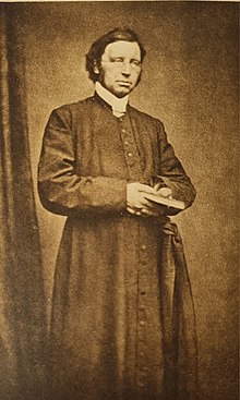 picture of a priest in cassock wearing glasses, standing holding an open book