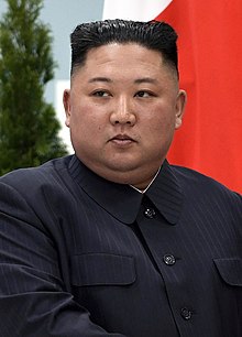 An official image of Kim Jong Un. He is wearing a black striped suit and standing in front of a flag.