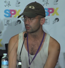 A man with round glasses, short hair, a green baseball cap with two eyes on it, and a white tank top speaks into a microphone while sitting.