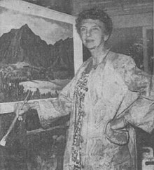 Portrait of a woman artist in an artist's smock with a large paint brush in her hand standing in front of an easel on which a painting of a mountain stands