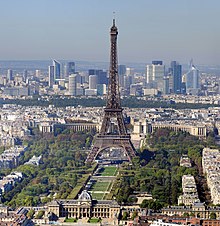 Farbige Obersicht vom Eiffelturm mit der langen Rasenfläche und dem École militaire im Vordergrund. An beiden Seiten sind Baumflächen und hinter dem Eiffelturm ist das Palais de Chaillot mit vielen Häusern. Im Hintergrund sind die Hochhäuser von La Défense.