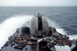 A large cannon on the front of a warship surrounded by empty shells; waves are breaking over the ship.