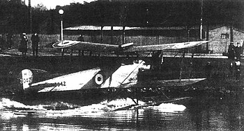 Three-quarter rear view of biplane on floats, taxiing along a stretch of water towards shore