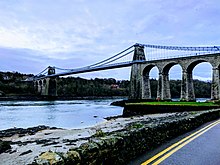 Pale blue metal suspension bridge carrying road traffic across a body of water with large stone arches at one end