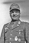 A man wearing a field cap and military uniform with various military decorations including an Iron Cross displayed at the front of his uniform collar.