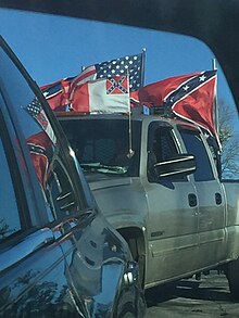 A large pickup truck has three clearly visible flags mounted onto it. The modern US flag, the Confederate Battle flag, and a Confederate National flag.