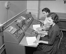 Black and white photo of two women sitting at desks with a punch machines