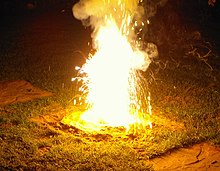 Ein Foto von einer weißen Flamme, aus der Rauch aufsteigt und leicht bläuliche, ansonsten ebenfalls weiße Funken fliegen. Die Flamme steigt aus einem flachen Kegel aus Erde auf, ist etwa 30 Zentimeter hoch und beleuchtet das Gras rund um den Kegel.