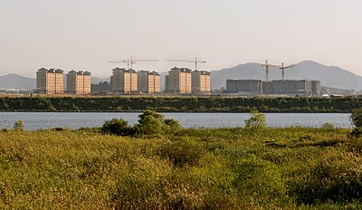 A photograph of the Camp Humphreys military base in South Korea on the other side of a river, showing six buildings of seven-storey dorm barracks on the left, and shorter wider buildings under construction on the right. Five cranes complete the skyline with a forested mountain range in the distance.