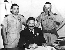 Half-length portrait of three military men behind a desk, all with pilot's wings on left breast pocket. One of the men, seated, has a large dark moustache and is wearing a dark winter uniform. The other two, standing on either side of the seated figure, wear short-sleeved tropical uniforms; one of them has a small moustache, the other has a holster on his belt and is smoking a pipe