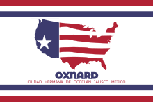 Three stripes, red, white, and blue from top to bottom, line the top and bottom of a white flag. Red and white stripes fill an outline of the contiguous United States in the center, while the left third is blue with a single white star in the lower half. Text in blue below the outline reads, "Oxnard," and small red text below that reads "CIUDAD HERMANA DE OCOTLAN JALISCO MEXICO."