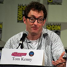 Tom Kenny, a tall White man with brown, curly hair and glasses, seats at a microphone looking off to his left