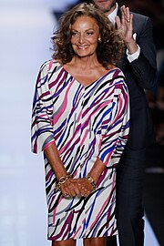 A woman with wavy and curly hair wearing a white dress with multi-colored stripes smiles on a fashion runway