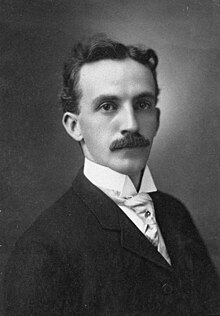 Black and white portrait of a man with short hair and a moustache wearing a dark suit with a high-collared shirt