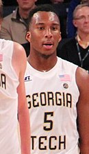 Waist high view of man with short black hair wearing a white Georgia Tech uniform, standing in front of the crowd, slightly obscured by another player at left