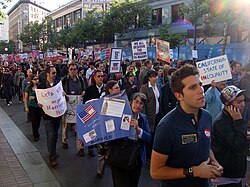 People peacefully marching up a street. Several are carrying signs displaying pro-same-sex marriage slogans, such as "We all deserve the freedom to marry." Most people have sad or serious facial expressions.