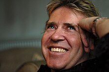 A headshot of Steve Lillywhite smiling, while his hand is pressed against the side of his face