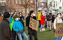 Das Bild zeigt eine Gruppe von Menschen. Zentral steht ein junger Mann mit schwarzer Mütze und weißer Atemschutzmaske, der ein Schild mit der Aufschrift „Fight for 1.5“ hält.
