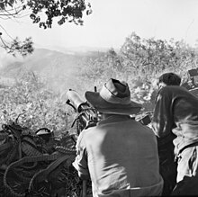 Soldiers firing a machine-gun towards a hillside in the distance. A large number of discarded ammunition belts are visible.