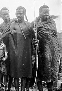 Black-and-white photo of three Maasai men in shúkà, two plain and probably red, one tartan.