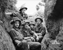 Five young Caucasian men in uniform wearing helmets sit below ground in a trench, facing the camera. The trench is central to the photograph and runs away from it, with the edges of the earthworks on each side. In the background, the skyline can be seen to the rear of the men, framed by the edges of the trench, which are covered in roots, grass, and other vegetation.