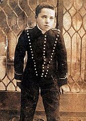 Schwarzweißfotografie eines kleinen Jungen, der bis zu den Knien porträtiert ist. Er blickt ernsthaft in die Kamera und trägt ein langärmliges Oberteil mit drei vertikalen Knopfreihen. Auf den Schultern und an den Ärmelenden sind Kordelverzierungen. Im Hintergrund ist eine Glaswand mit Sprossenverzierungen.