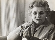 A sepia photo of a middle-aged woman in a short-sleeve collared shirt sitting and gazing at an item she is holding.