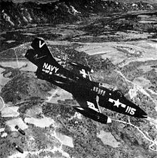 A single-seat jet aircraft with US Navy markings in mid-air, flying in profile from left to right in a downward angle of attack below the camera. To the bottom left, two bombs are falling away from the aircraft towards an unseen target in the fields below.