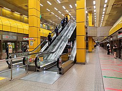 Underground island platform with escalators between two yellow pillars leading up to the concourse