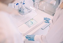 Eine Person im weißen Kittel und mit blauen Gummihandschuhen steht vor einem Labortisch, auf dem ein 3D-Bioprinter steht. Dieser besteht aus einer vertikal ausgerichteten Spritze, Linearantrieben und einer Petrischale