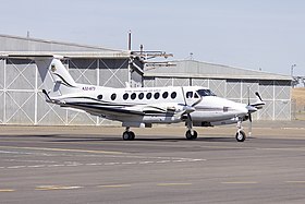 Colour photograph of a white aircraft on the tarmac of an airport in front of a grey building