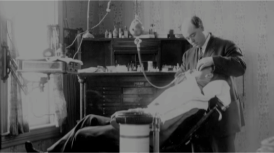 Black and white photograph of a small, regular, room, a window to the left with a dental chair facing this direction. The dentist stands behind the chair to the right, with dental paraphernalia occupying many surfaces: an oxygen tank is front and center, a tall table is laid with tools in front of a wardrobe in the rear of the image, and a drawer is hung by the window with more tools.