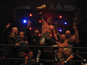 Ten members of the Bullet Club group celebrate in a professional wrestling ring. Several are using a hand gesture, extending the index and little finger, with the tips of the renaming fingers touching.