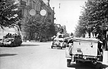 Frontale Schwarzweißfotografie von einer Straße mit Militärfahrzeugen. Links und rechts sind Baumalleen. Links steht ein breites, altes Gebäude.