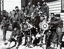 A group of twelve uniformed US Army servicemen gathered around two Native American men dressed in traditional tribal clothing