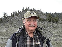 L. David Mech wearing an unzipped coat over a checkered shirt, and a khaki-colored cap with a Yellowstone logo, smiling and looking just left of camera