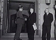 Black and white photograph of a man wearing military uniform shaking the hand of a man in a formal suit. The two men are standing in front of a stone gateway, and another man wearing a formal suit is looking on from the right of the image.
