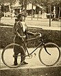 Eine junge Frau, die mit Stiefeln, Pluderhose, langer Jacke und Mütze bekleidet ist, steht auf einem Gehweg und hält ihr Herrenrad an Lenker und Sattel