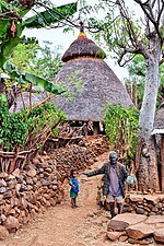 A child and an adult in a village with stone walls and a house with a thatched roof