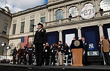 A woman in an American Navy uniform stands before a microphone with several people behind her on stage. The stage is in front of a building with a sign that reads "#27!".