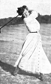 White woman outdoors in 1901, wearing long white dress, swinging golf club.