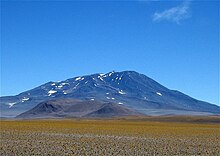 A mountain with snow patches rises above a smaller hill, which in turn rises above a plain covered with sparse yellow plants