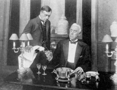 Two white, clean-shaven men: one is a butler, standing, pouring wine for the other, seated