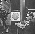 A sitting young man holds a microphone in his left hand while manipulating the console of an apparatus with his right. To his left a large television camera is trained on a large, circular cathode ray tube display.