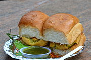 Vada pav served with a side of green chilli pepper, red peanut and garlic chutney, and green chutney.